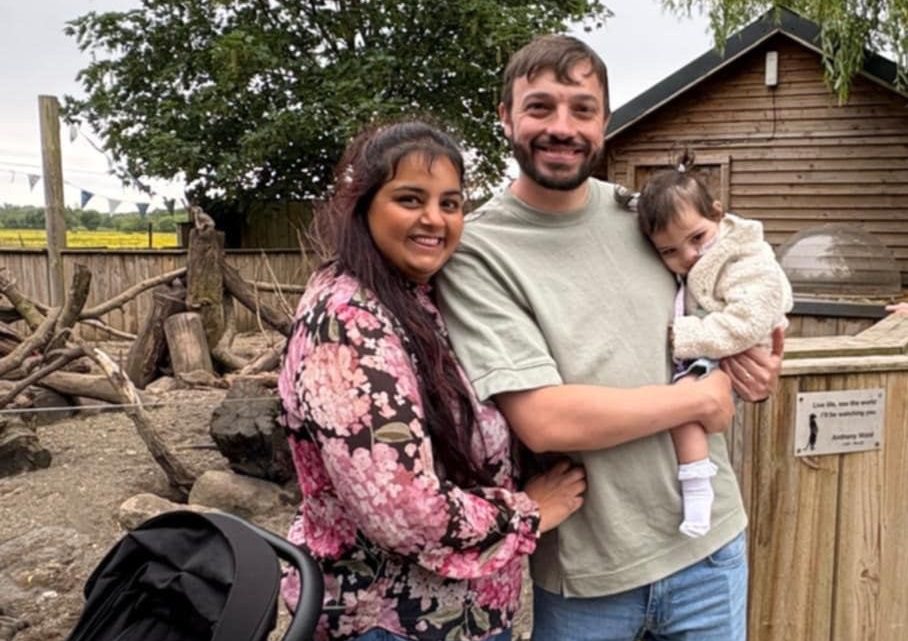 Woman and man holding a little girl at a zoo smiling at the camera. The little girl is asleep in her father's arms.