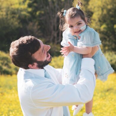 Man wearing a white shirt lifts a little girl wearing a blue dress into the air. The girl has a feeding tube into her nose and is laughing.