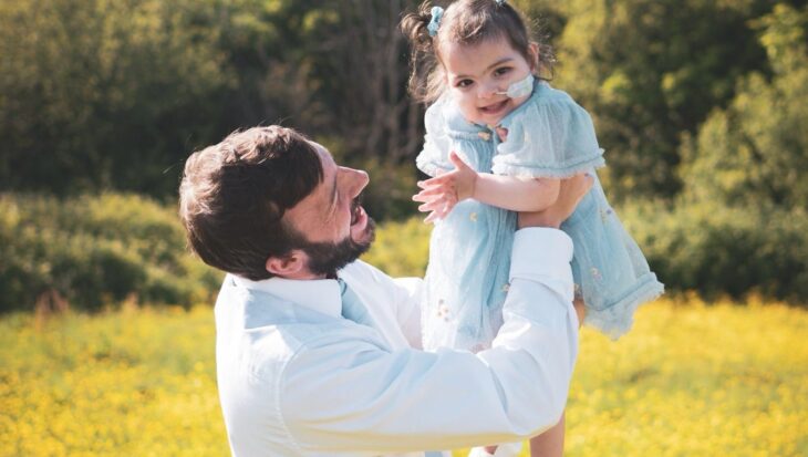 Man wearing a white shirt lifts a little girl wearing a blue dress into the air. The girl has a feeding tube into her nose and is laughing.
