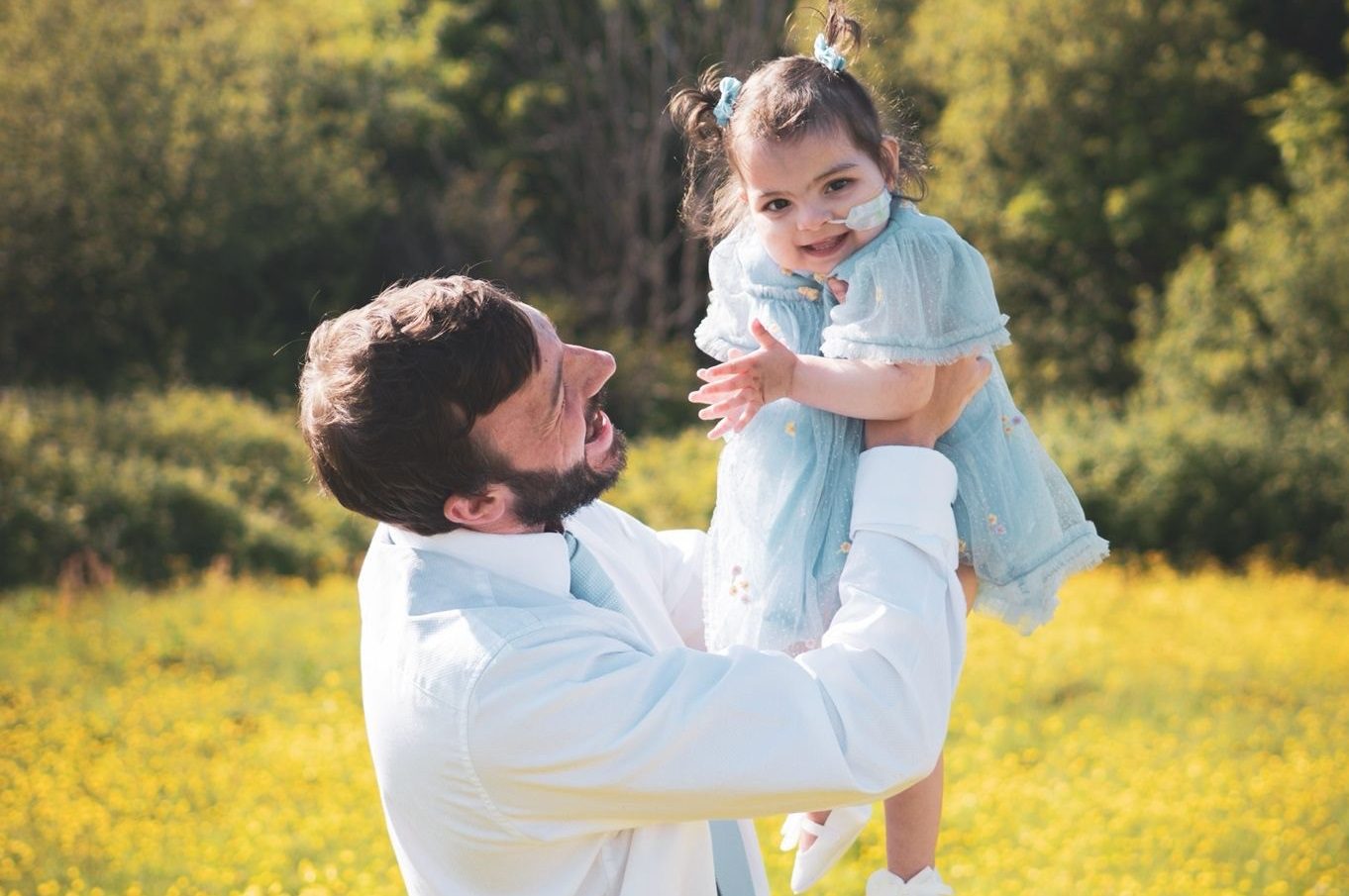 Man wearing a white shirt lifts a little girl wearing a blue dress into the air. The girl has a feeding tube into her nose and is laughing.