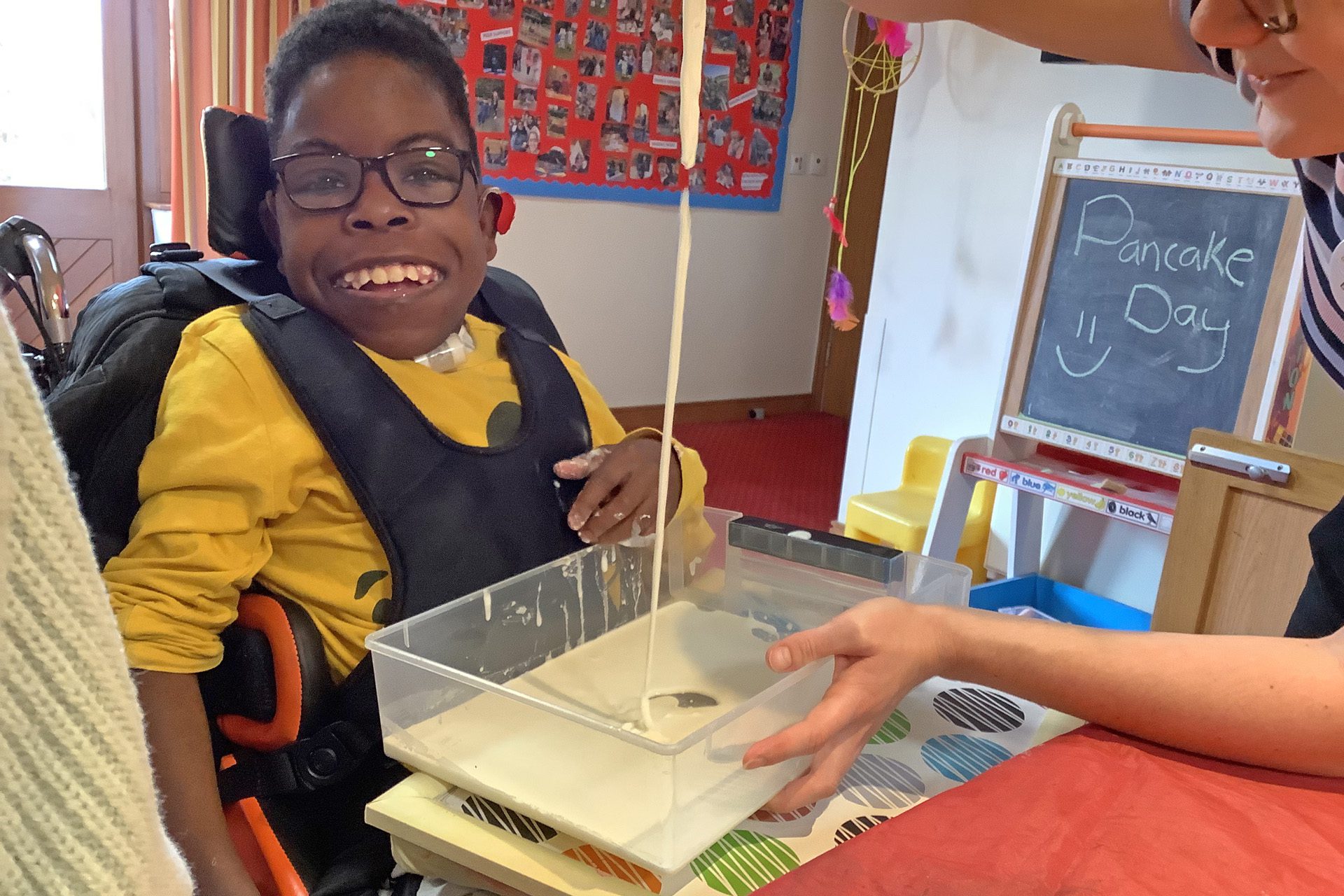 Young boy wearing a harness in a wheelchair smiling at the camera as a woman pours a stream of pancake batter into a plastic tub in front in a room with crafts and art on the walls.