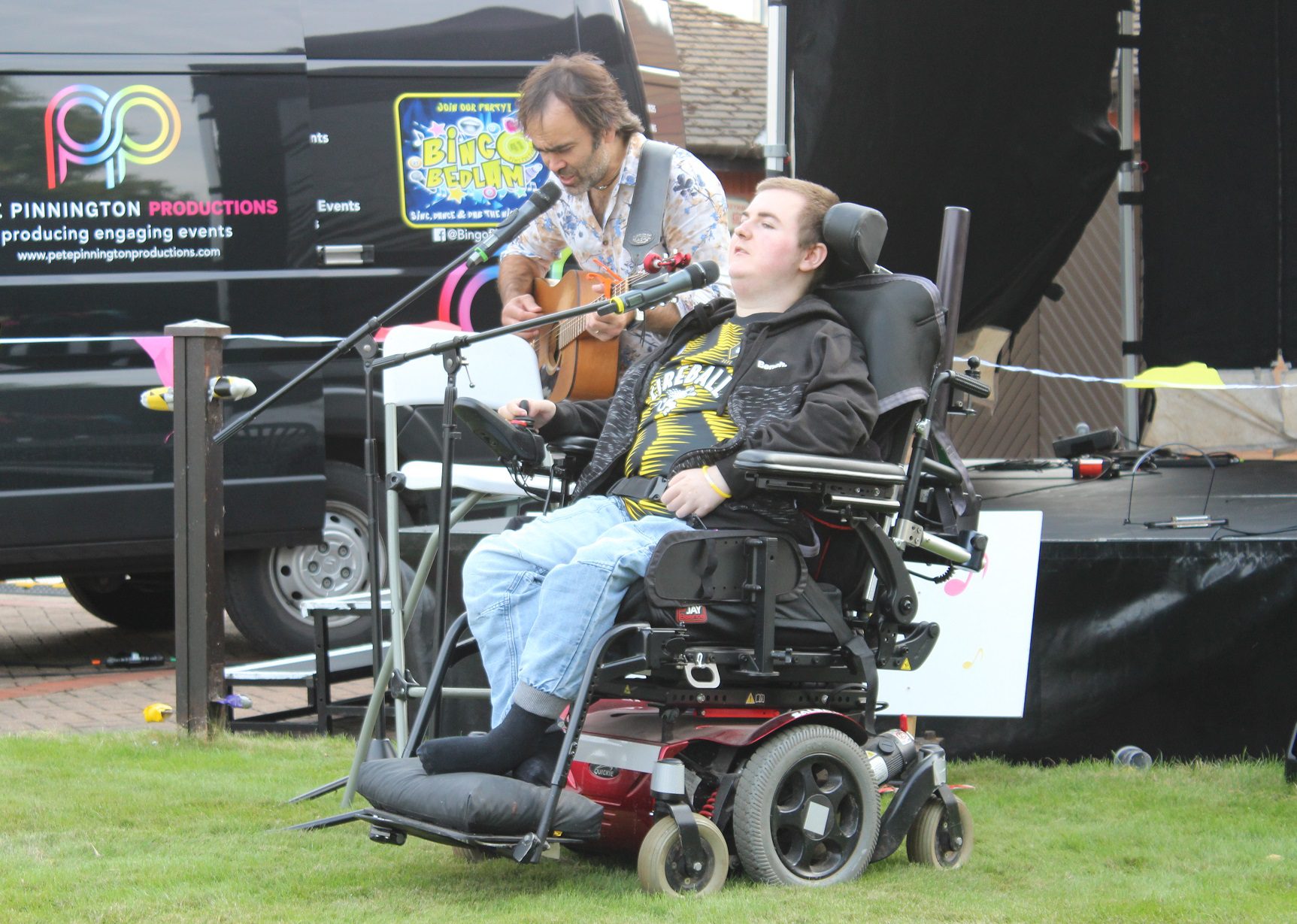 Young person in an electric wheelchair singing into a microphone alongside a man playing a guitar in front of a stage