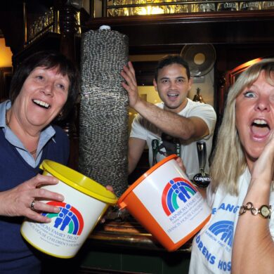 Two women holding charity buckets stood in a bar in front of a tower of ten pence coins and a man stood behind preparing to push the stack of coins over