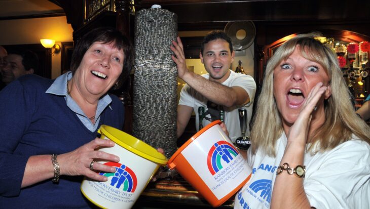 Two women holding charity buckets stood in a bar in front of a tower of ten pence coins and a man stood behind preparing to push the stack of coins over
