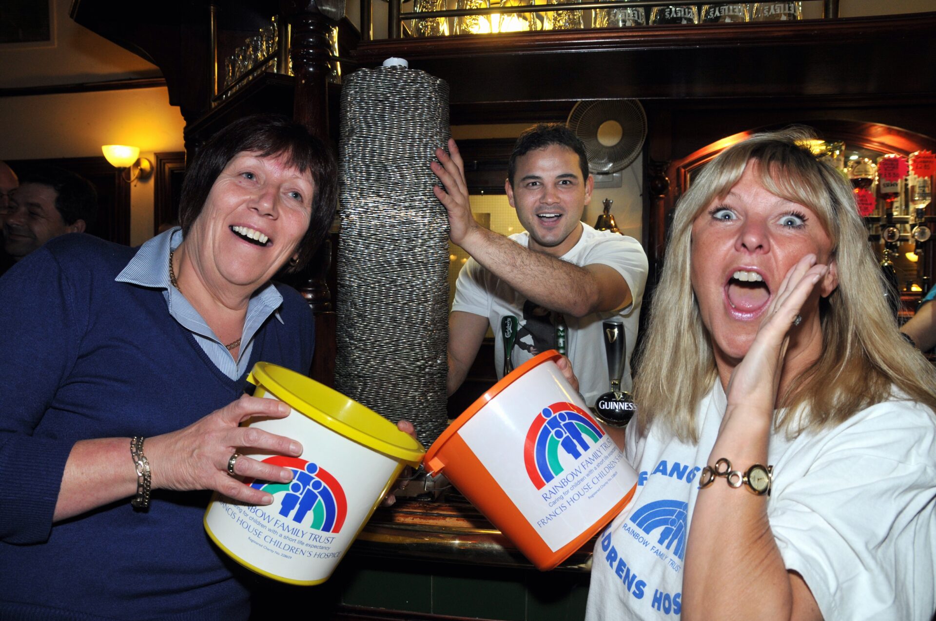 Two women holding charity buckets stood in a bar in front of a tower of ten pence coins and a man stood behind preparing to push the stack of coins over