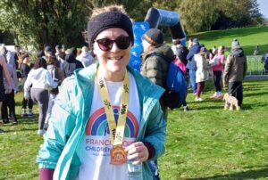 Woman wearing a black headband and a green jacket holding a running medal stood in front of spectators at the finish of a race.