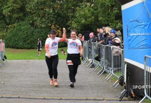 Two women wearing white t-shirts and black leggings holding hands in the air as they run towards the finish line of a race in a park.