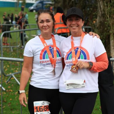 Two women wearing white t-shirts with running race medals around their necks stood in a park.