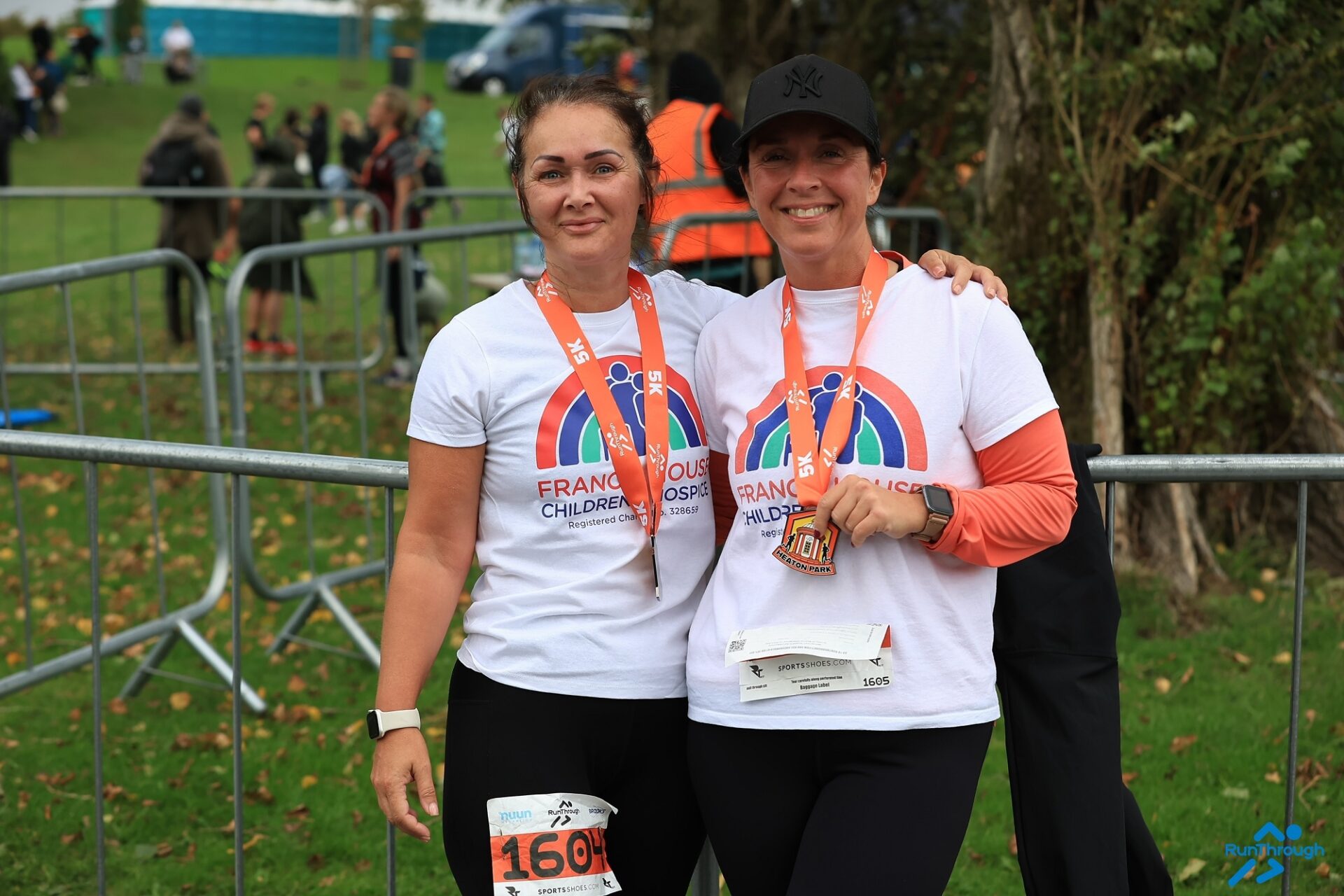Two women wearing white t-shirts with running race medals around their necks stood in a park.