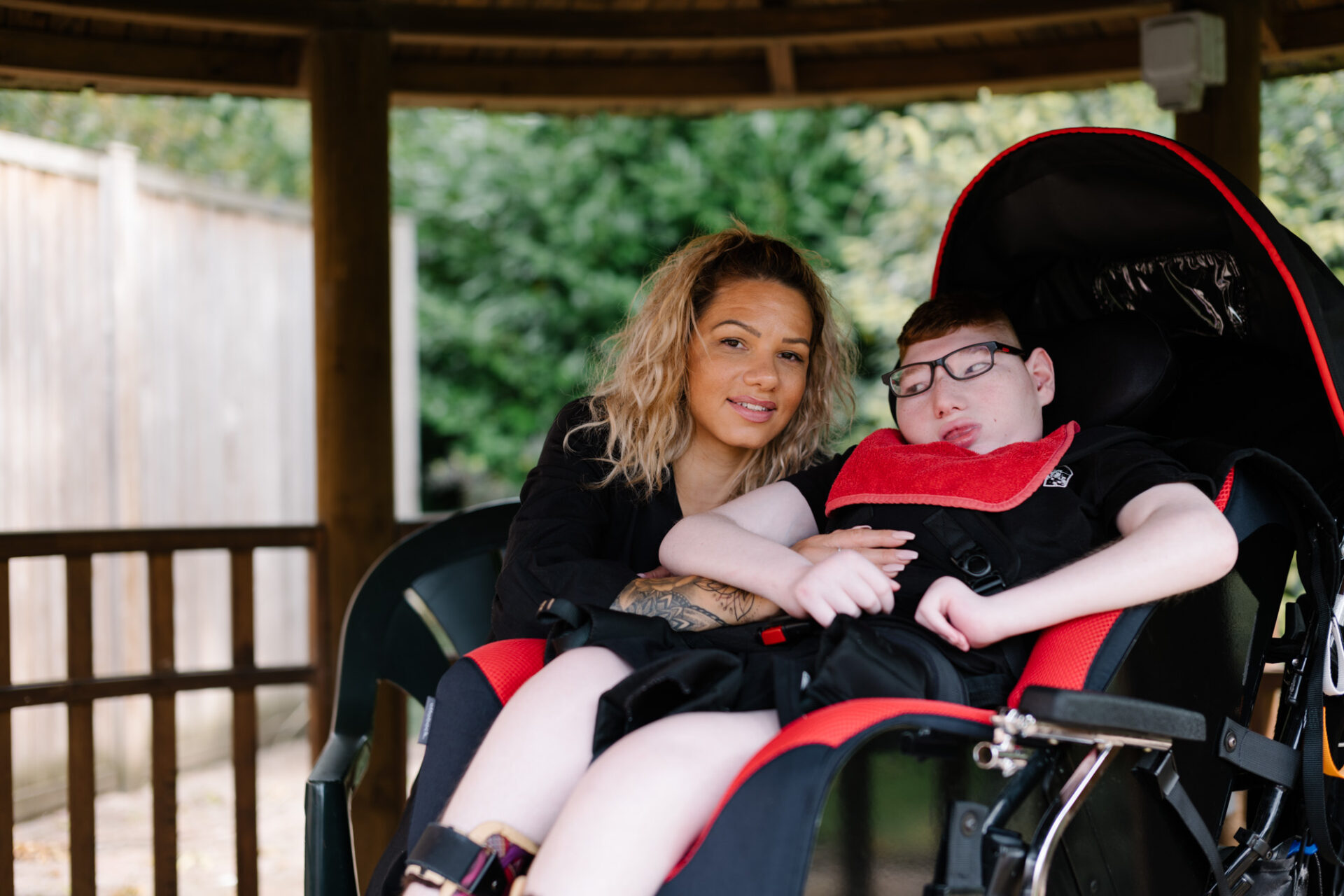 Young woman with curly blonde hair sat smiling at the camera next to a young boy seated in a wheelchair in a gazebo in a garden