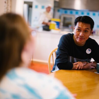 Man sat at a pine kitchen table with a cup of tea looking at a woman with long brown hair with her back to the camera out of focus.