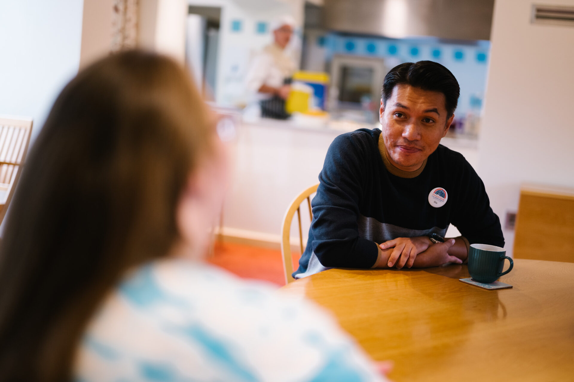 Man sat at a pine kitchen table with a cup of tea looking at a woman with long brown hair with her back to the camera out of focus.