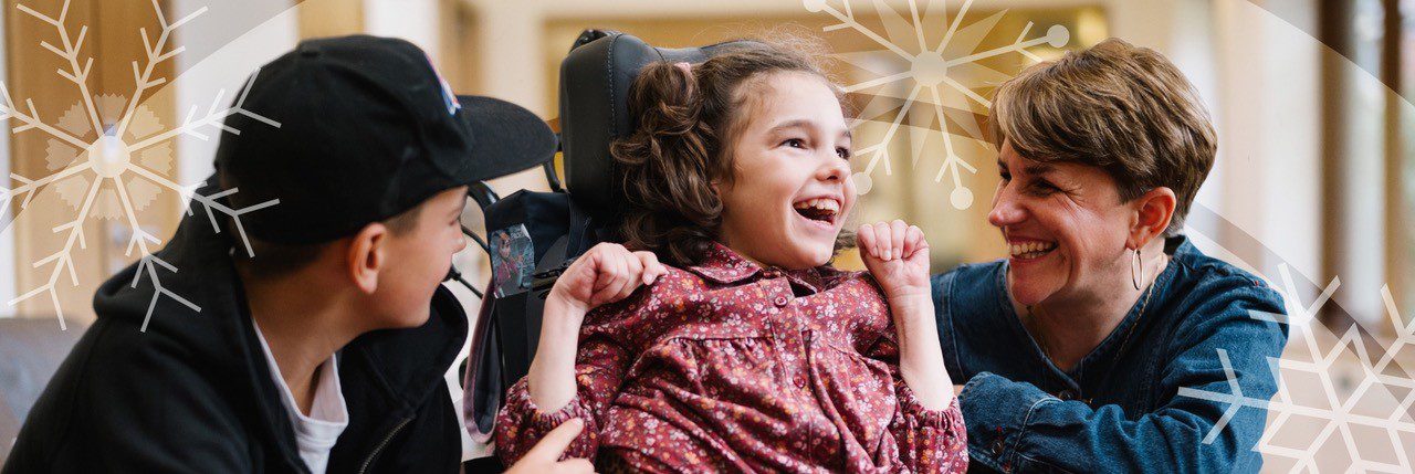 Young girl in a wheelchair wearing a pink dress sat between a young boy wearing a black baseball cap and a woman in a blue denim shirt looking and smiling at the girl.