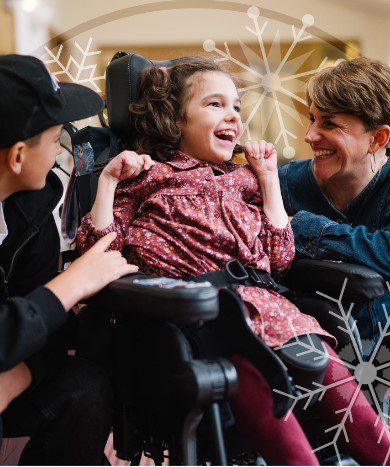Young girl in a wheelchair wearing a pink dress sat between a young boy wearing a black baseball cap and a woman in a blue denim shirt looking and smiling at the girl.