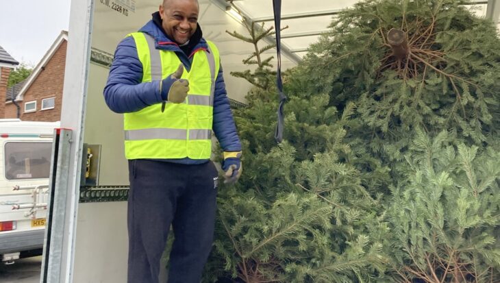 Man wearing a hi-vis jacket stood in the back of a white van containing Christmas trees to be taken for recycling