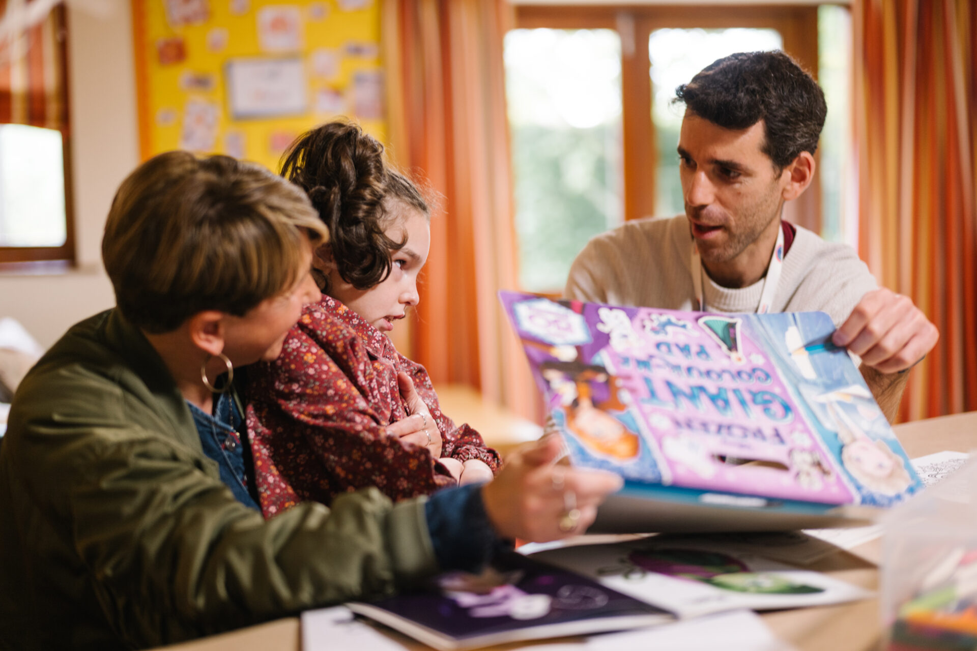 Man holding a child's reading book sat next to a young girl with brown hair and her mother wearing a green jacket in a play room with art on the walls