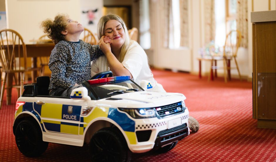 Young girl sat in a toy police car a woman with blonde hair holds the girl's hand against her cheek and smiles