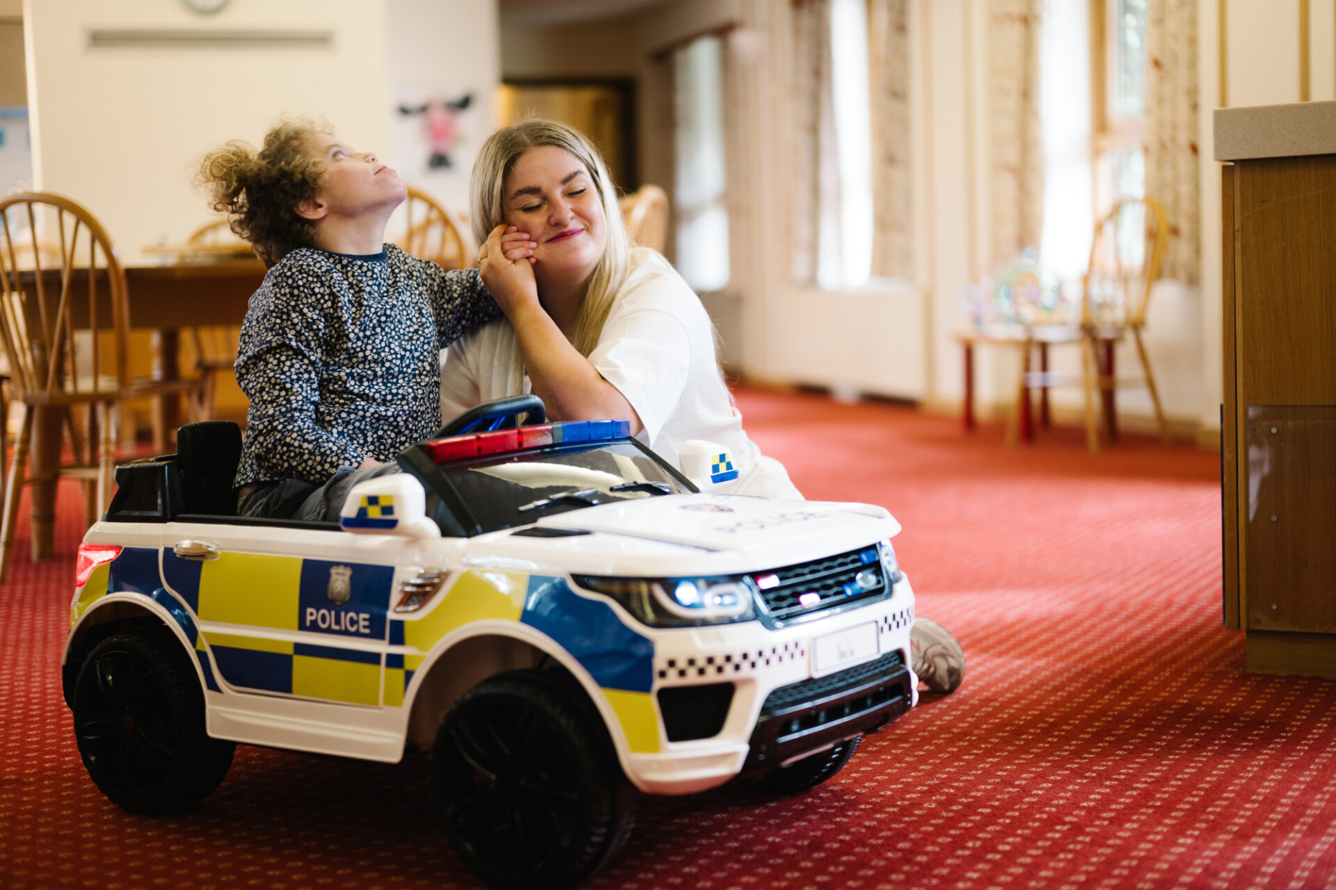 Young girl sat in a toy police car a woman with blonde hair holds the girl's hand against her cheek and smiles