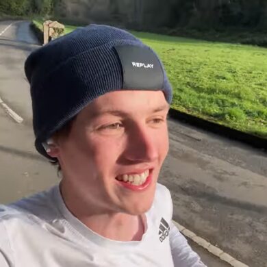 Man wearing a white top and black running hat as he runs along a road smiling
