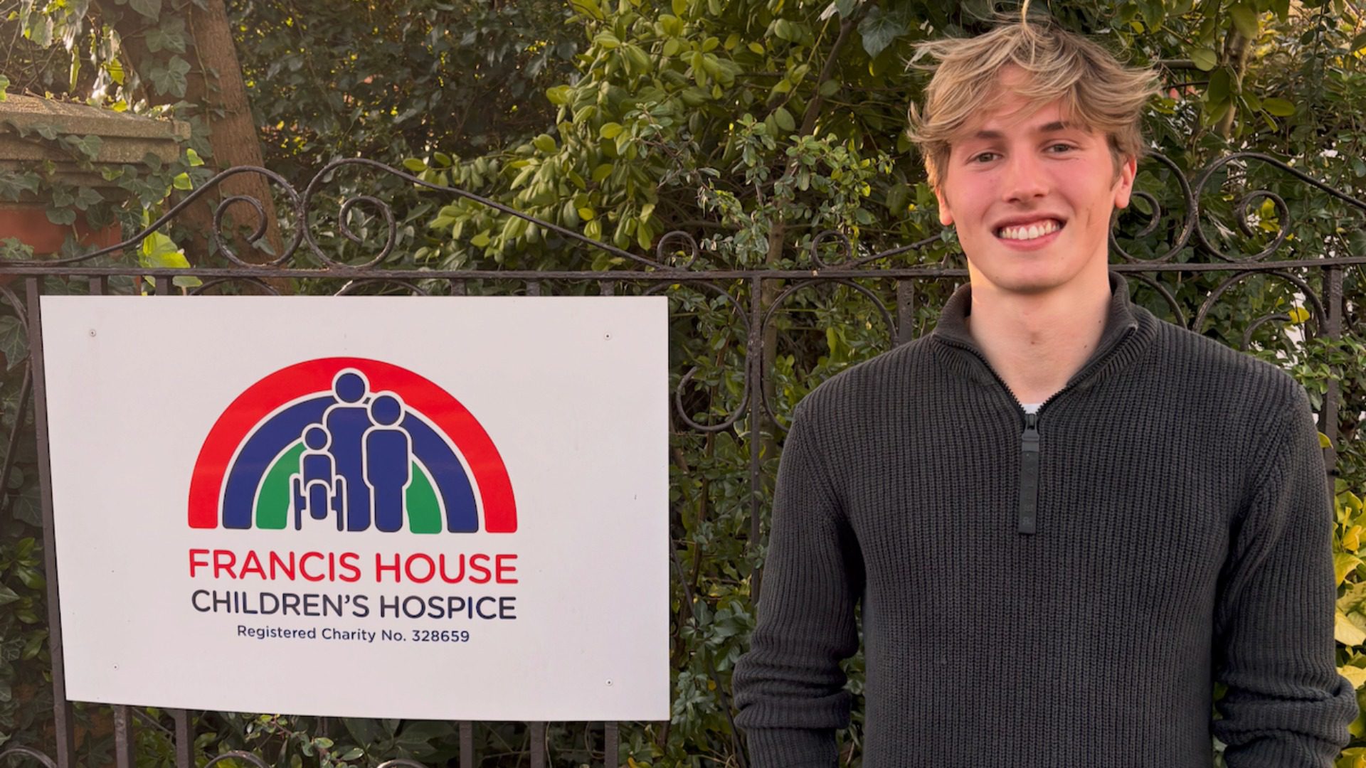 Man with blonde hair wearing a black top stood next to a sign on a gate which reads Francis House Children's Hospice