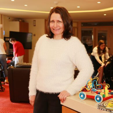 Woman wearing a white jumper stood in a lounge at a children's hospice with toys and people in the background