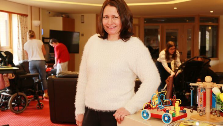 Woman wearing a white jumper stood in a lounge at a children's hospice with toys and people in the background