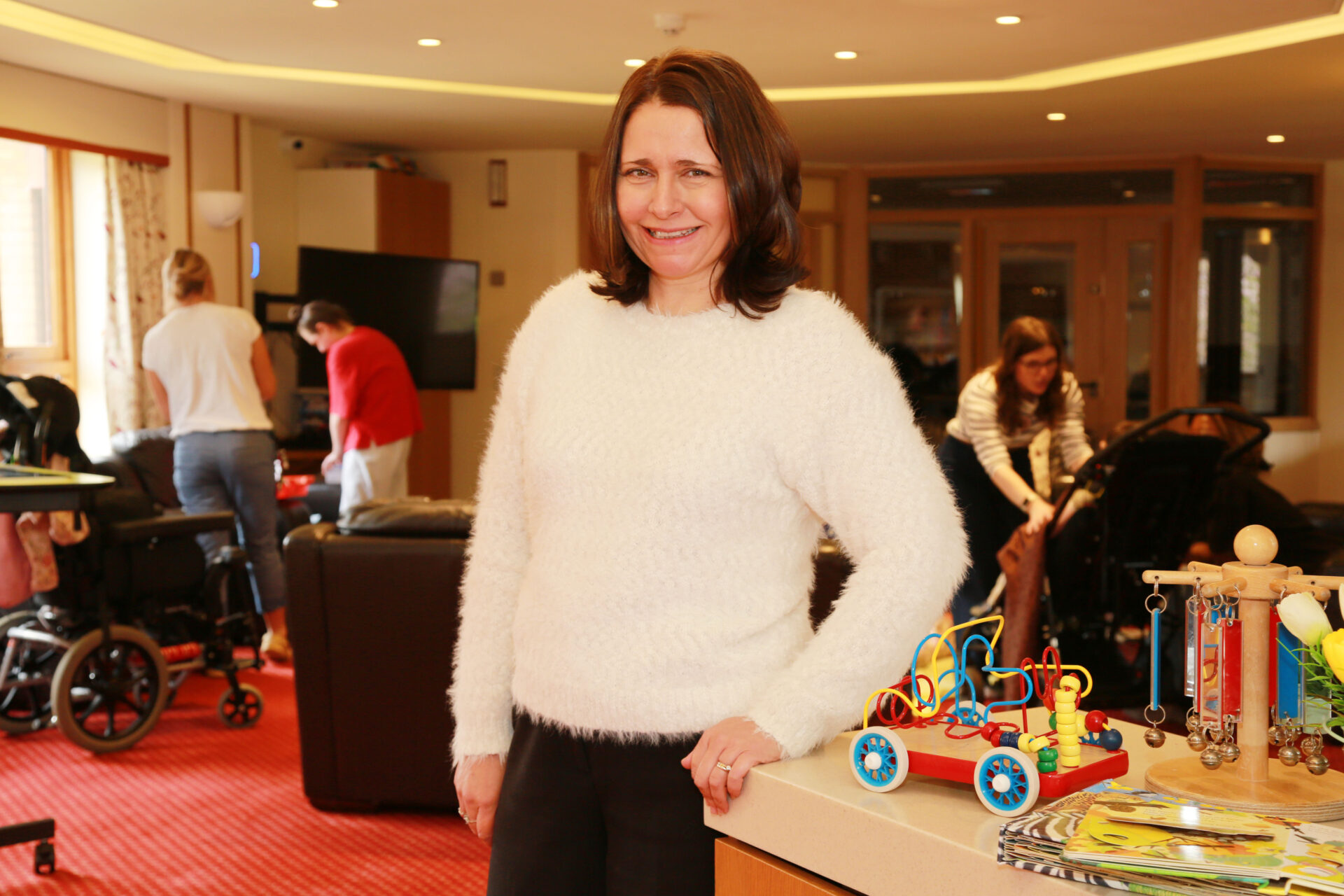 Woman wearing a white jumper stood in a lounge at a children's hospice with toys and people in the background