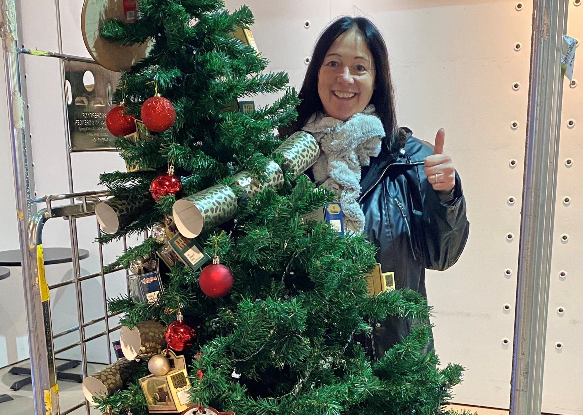 Woman giving a thumbs up and smiling at the camera stood next to a Christmas tree with crackers and baubles