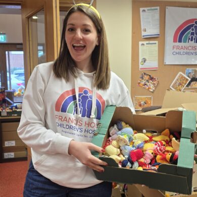 Young woman with long brown hair wearing a white hoodie holding a box of multi coloured knitted chicks stood in an office setting.