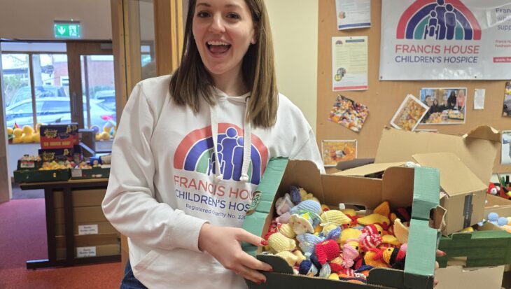 Young woman with long brown hair wearing a white hoodie holding a box of multi coloured knitted chicks stood in an office setting.