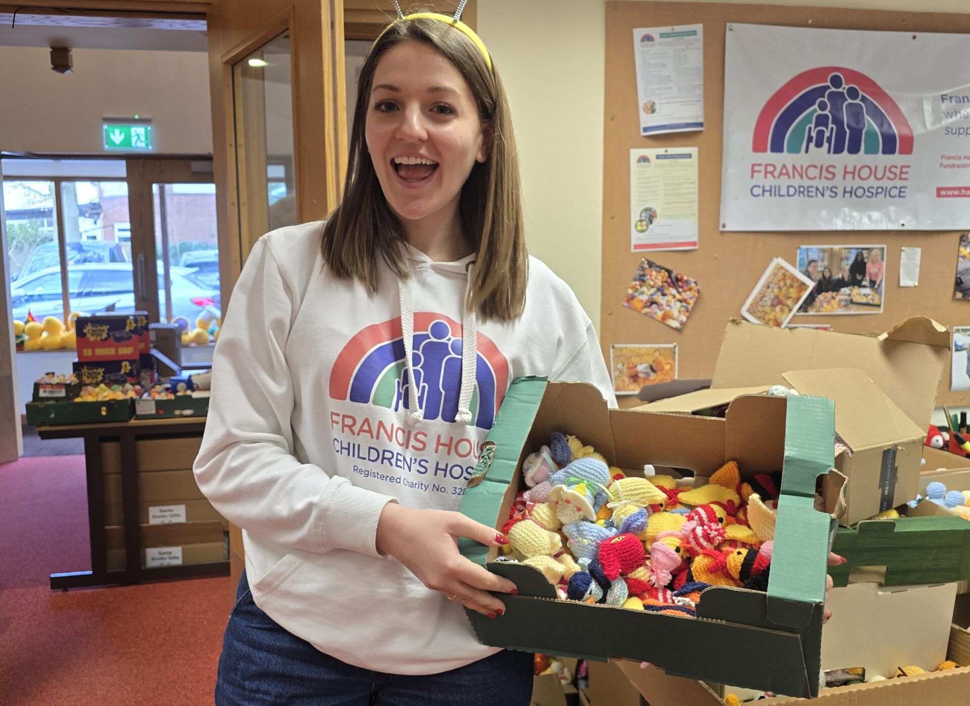 Young woman with long brown hair wearing a white hoodie holding a box of multi coloured knitted chicks stood in an office setting.
