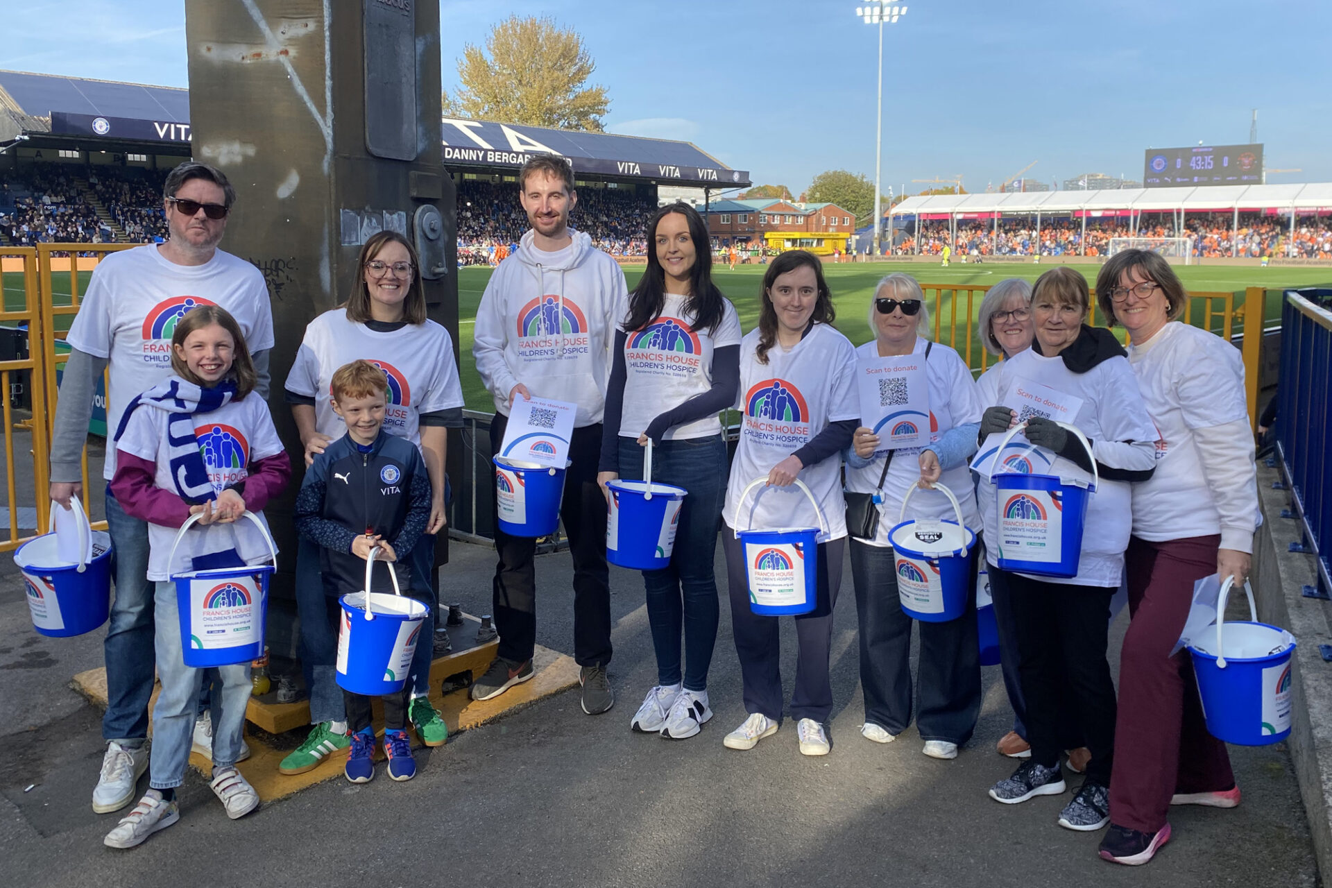 Row of people wearing white charity t-shirts and holding blue plastic buckets stood at a football ground