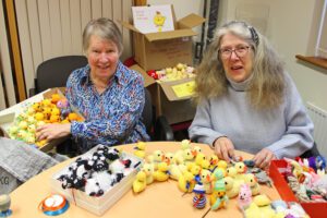 Two women holding boxes of knitted chicks sat behind a table
