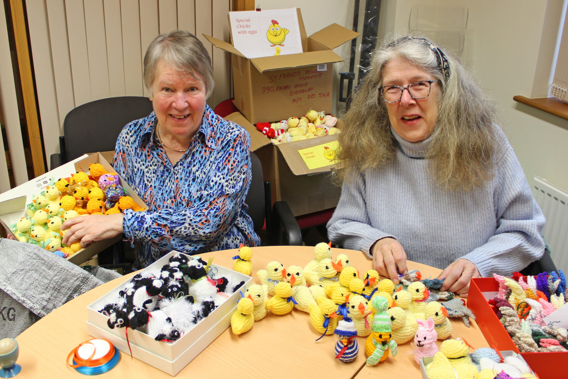 Two women holding boxes of knitted chicks sat behind a table