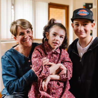 Woman wearing a blue top holding a young girl dressed in a pink dress sat alongside a young boy wearing a black cap and top.