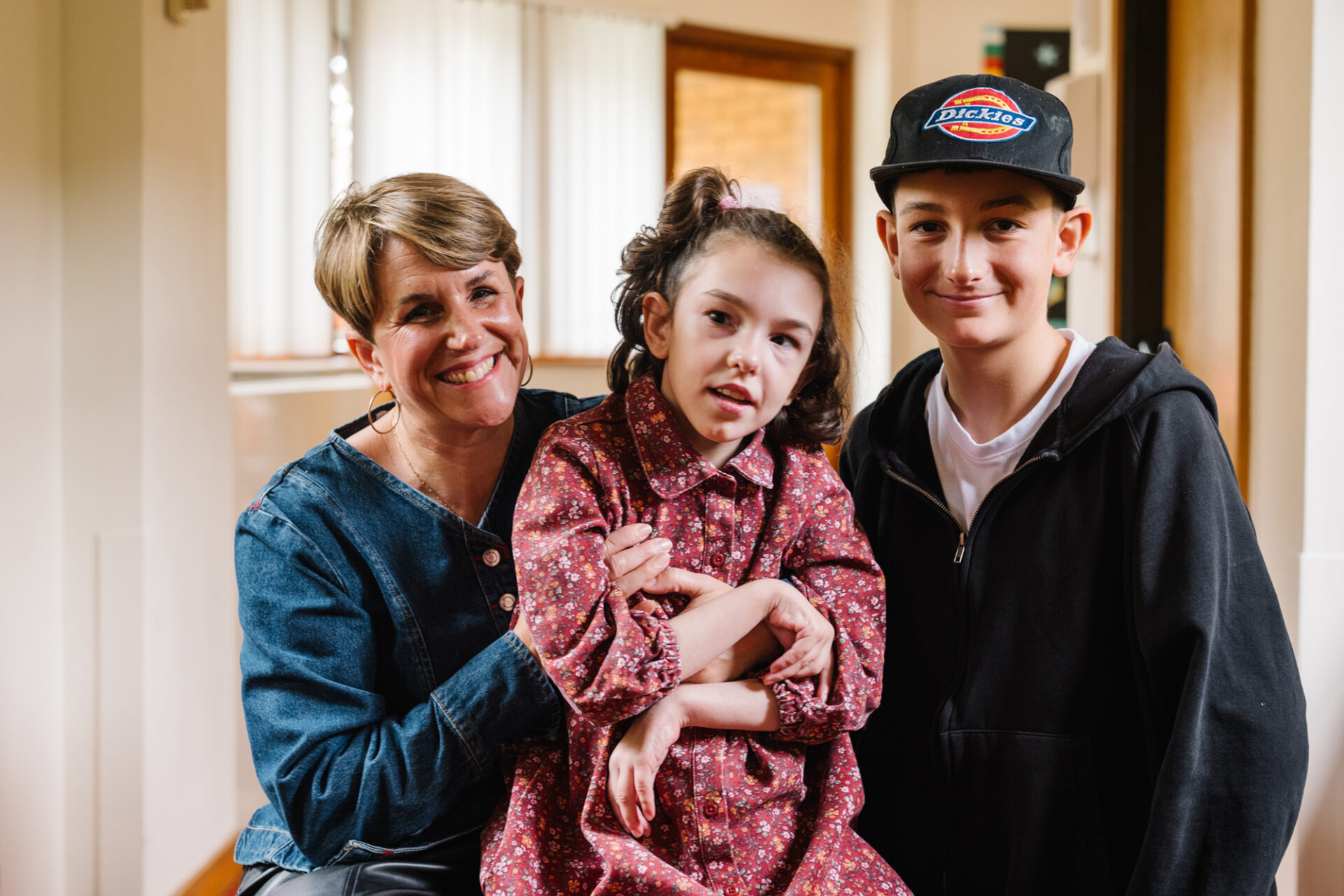 Woman wearing a blue top holding a young girl dressed in a pink dress sat alongside a young boy wearing a black cap and top.