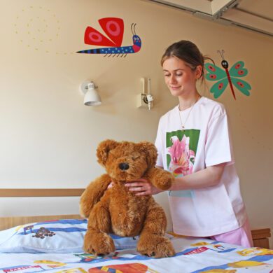 Young woman wearing a white t-shirt places a orange teddy bear on a bed in a child's bedroom with painted bug characters on the walls