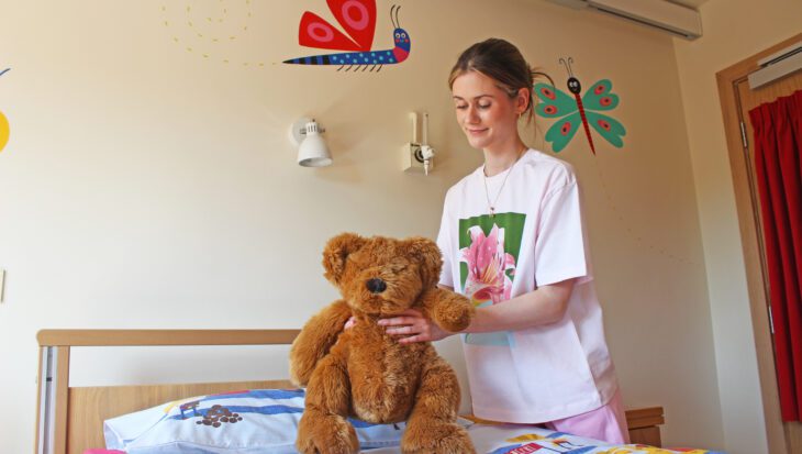 Young woman wearing a white t-shirt places a orange teddy bear on a bed in a child's bedroom with painted bug characters on the walls