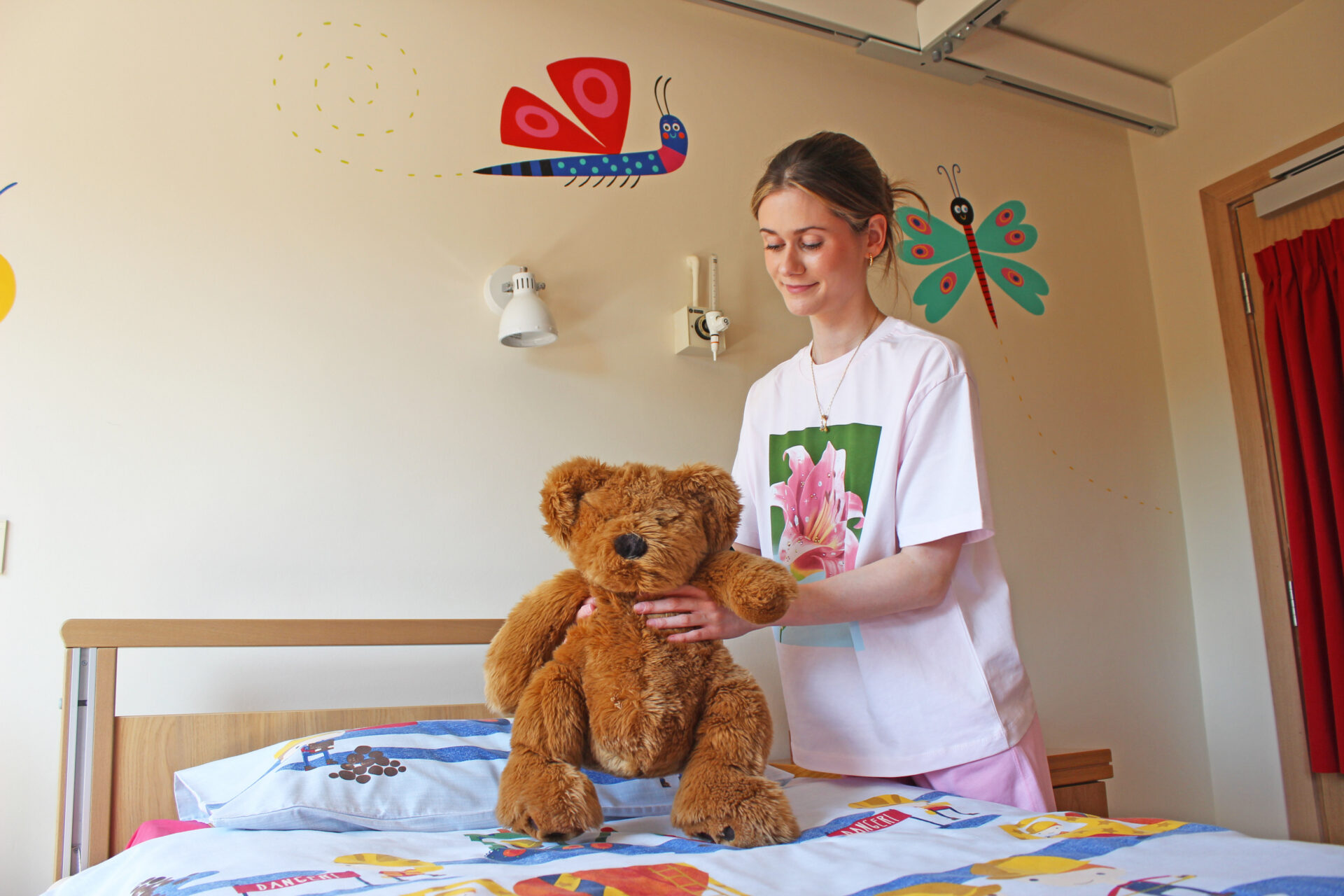 Young woman wearing a white t-shirt places a orange teddy bear on a bed in a child's bedroom with painted bug characters on the walls