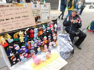 Man sat in a high street next to a display of knitted dolls for sale to charity
