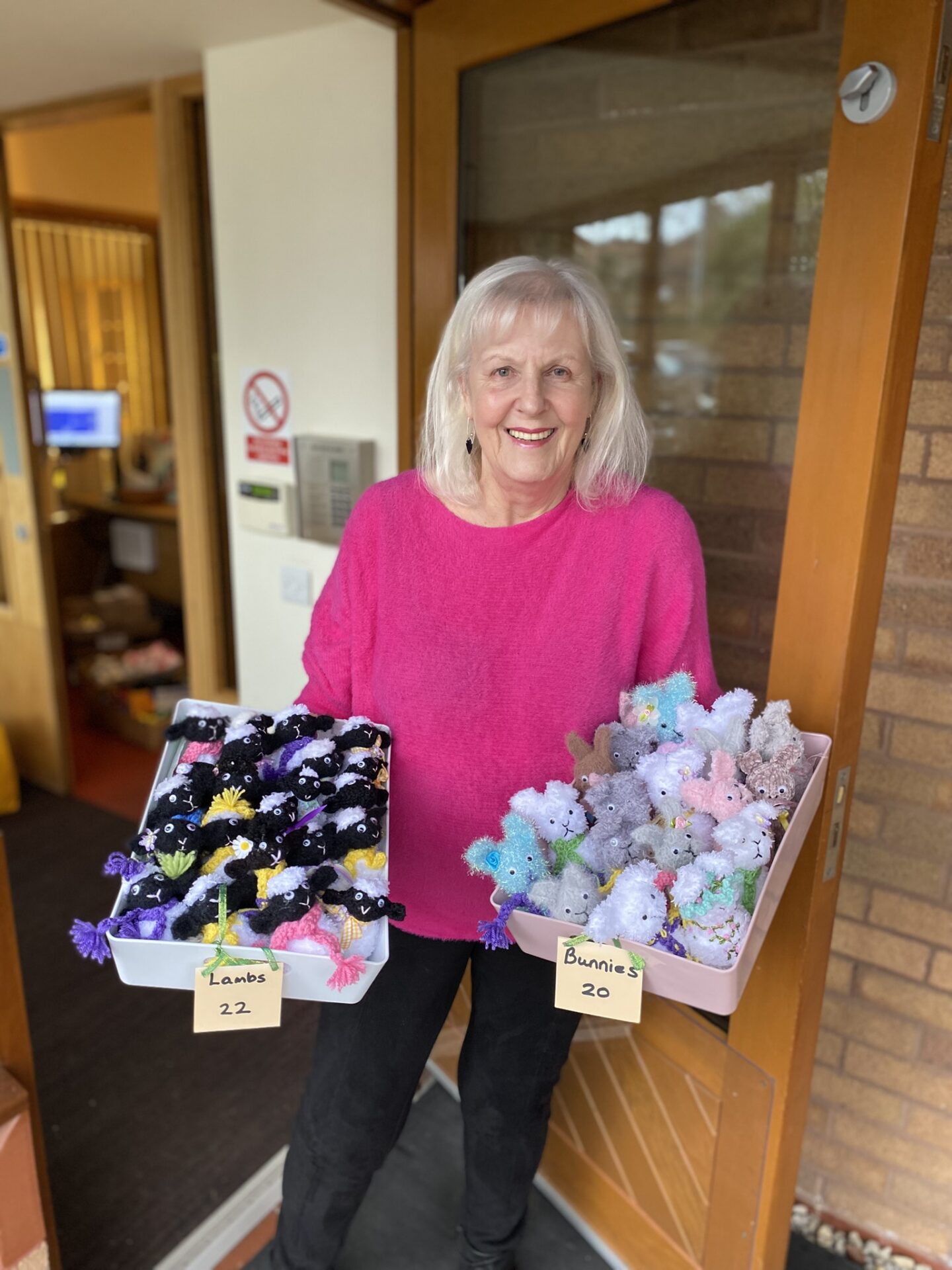 A woman stands outside Francis House holding a box in each hand, one containing lots of knitted lambs and the other knitted bunnies