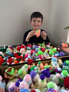 A young boy sits in front of lots of knitted chicks whilst demonstrating how to fit an egg inside a chick