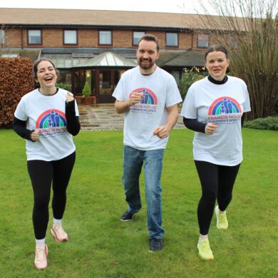 Two women and one man wearing running t-shirts running on a grassy lawn in front of a brick building