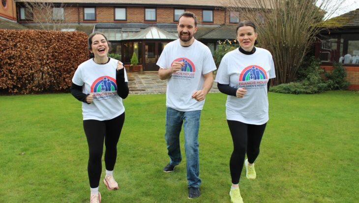 Two women and one man wearing running t-shirts running on a grassy lawn in front of a brick building