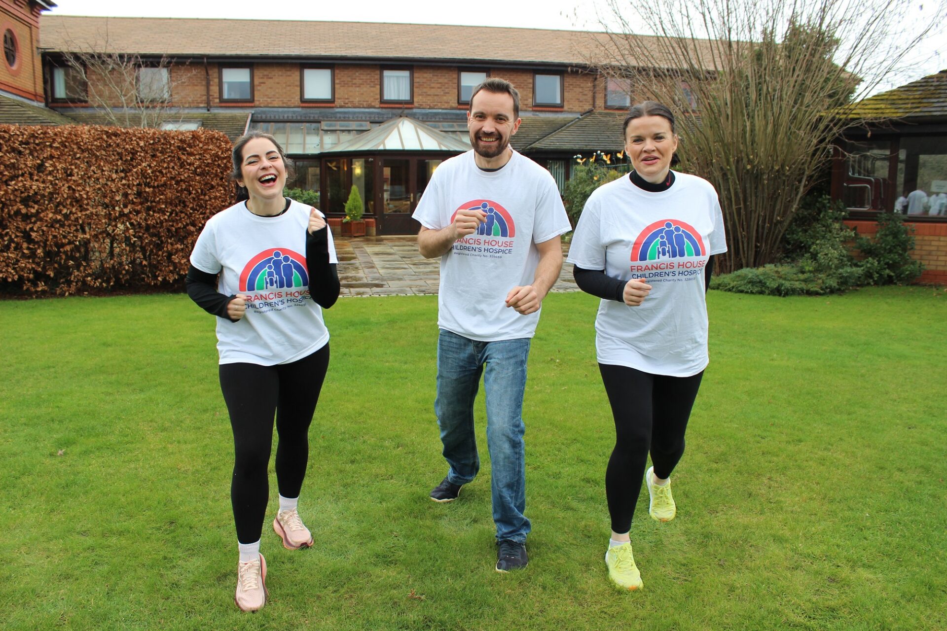 Two women and one man wearing running t-shirts running on a grassy lawn in front of a brick building