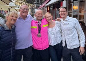 Group of five men and women with their arms around each other smiling at the camera. The man in the middle is wearing a bright pink running top and has a London Marathon medal around his neck.