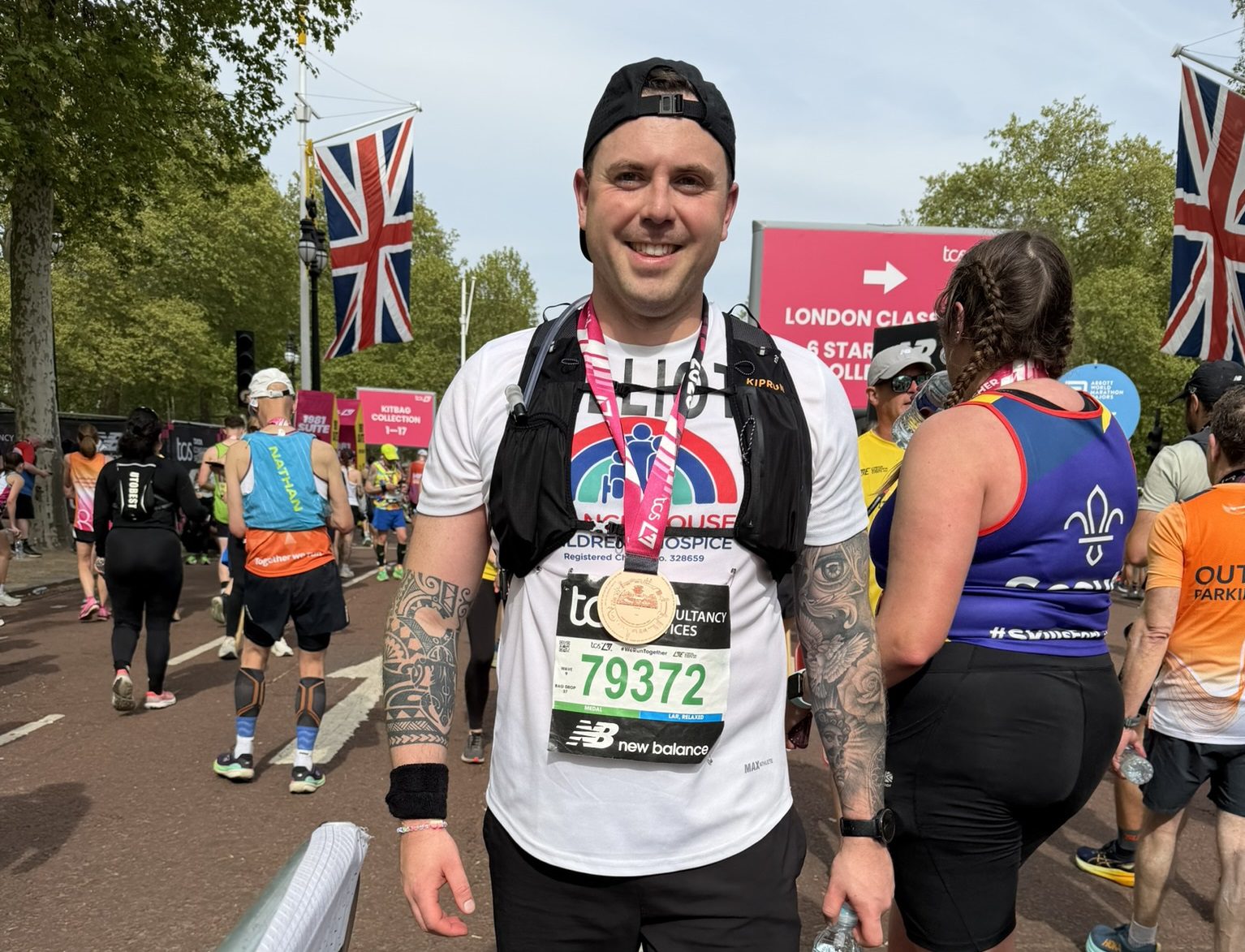 Man stood on a road with other runners wearing a London Marathon medal around his neck smiling at the camera