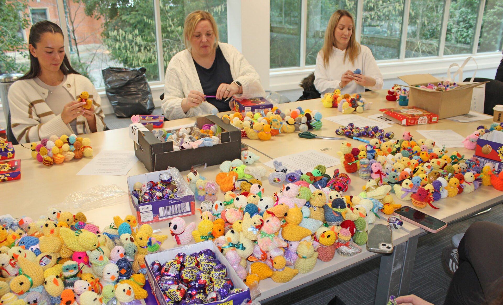 Three women sat around a large table in an office stuffing creme eggs into knitted chicks