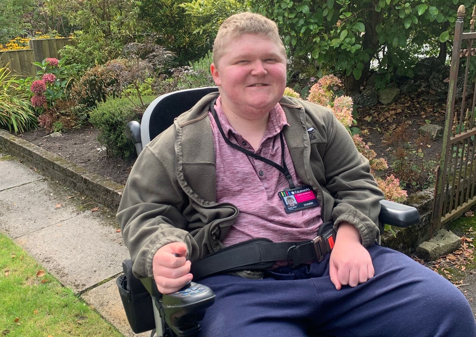 Young man with blonde hair smiling at the camera. He is seated in a wheelchair in a garden.