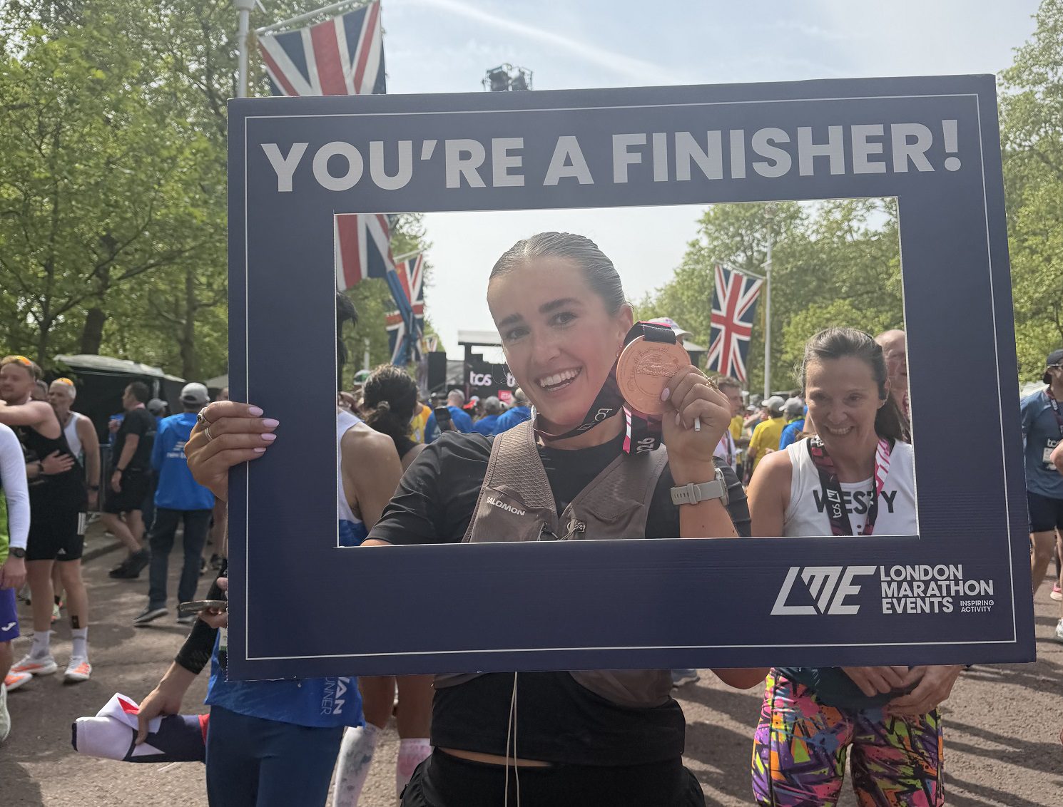 Woman holding a London Marathon medal and sign that says You're a finisher! she stands on a road with other runners.